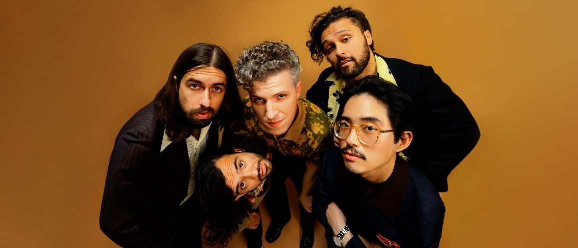 Gang of Youths, band of five men huddled together, looking into the camera with an organ backdrop behind them.
