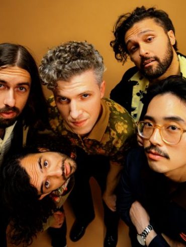 Gang of Youths, band of five men huddled together, looking into the camera with an organ backdrop behind them.