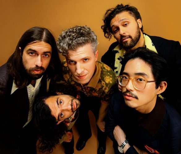 Gang of Youths, band of five men huddled together, looking into the camera with an organ backdrop behind them.