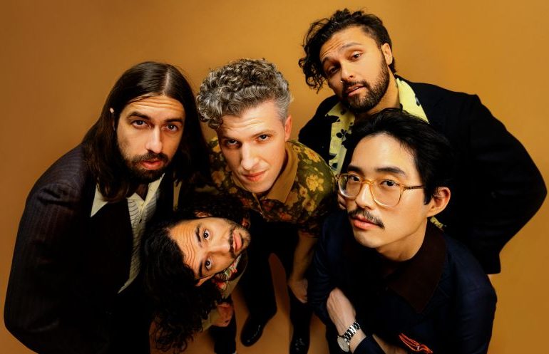 Gang of Youths, band of five men huddled together, looking into the camera with an organ backdrop behind them.